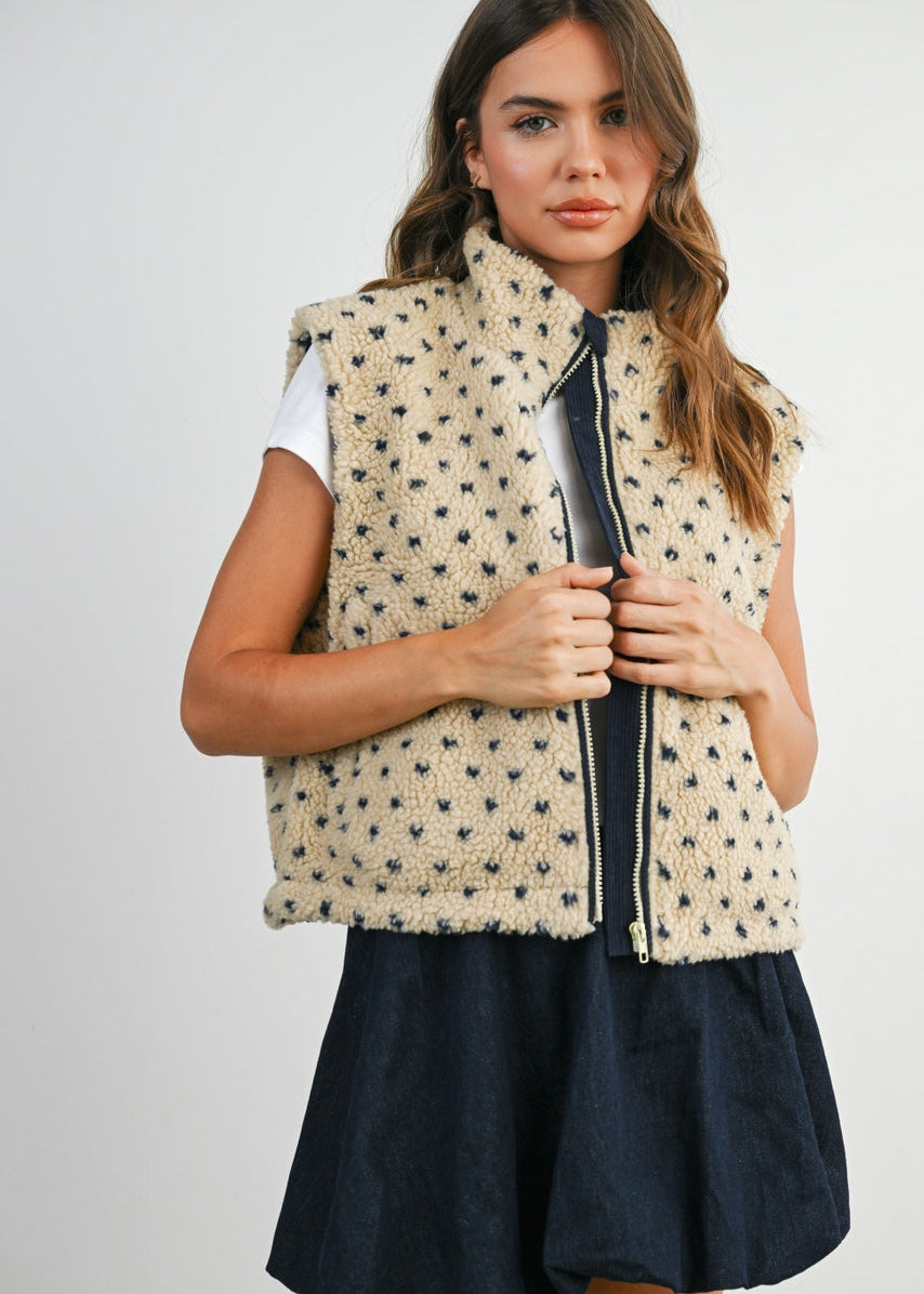 Woman wearing a patterned vest over a white shirt with a navy skirt on a plain background