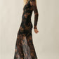 A woman modeling a long-sleeve black sheer mesh maxi dress with a brown floral pattern, paired with brown studded heels, against a plain beige studio background.