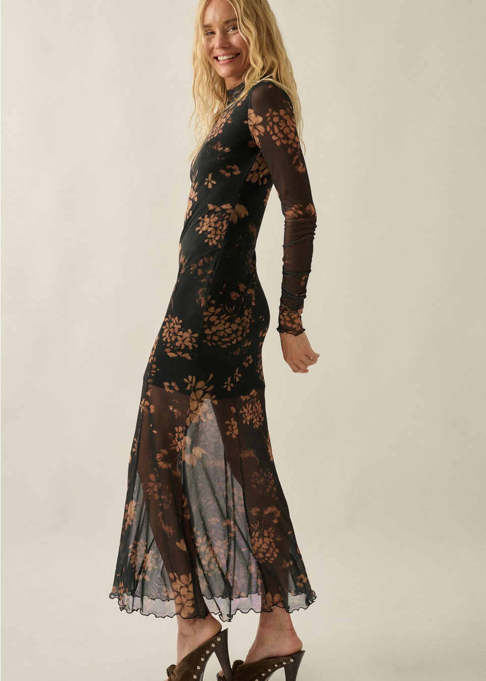 A woman modeling a long-sleeve black sheer mesh maxi dress with a brown floral pattern, paired with brown studded heels, against a plain beige studio background.